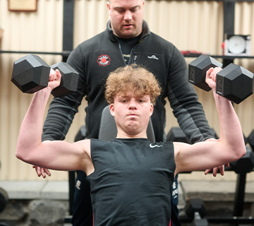 A student doing a dumbbell shoulder press with a coach behind him